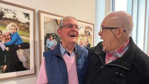 Two white gentlemen in their 80's laugh together, standing in front of photos of one of the men and their family