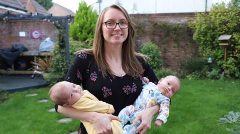 Emily in a garden holding her newborn twins in each arm