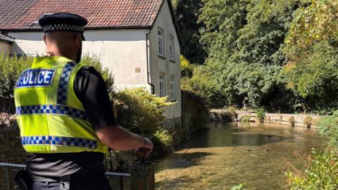 A police officer walking on a raised footpath alongside a shallow, clear river. On the opposite side is a white rendered cottage with a chimney and red tiled roof.
