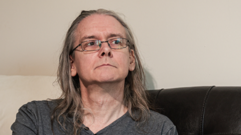 Martin Atkinson, a middle-aged man with long grey hair, sits on a bed looking to the side of the camera. He wears black jeans and a long-sleeved grey shirt. 
