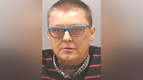 Mugshot of Heslop. She has very short dark shaved hair and blue framed glasses, with a tattoo of flowers across her throat.