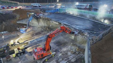 Two cranes work on destroying a bridge that runs over the M25