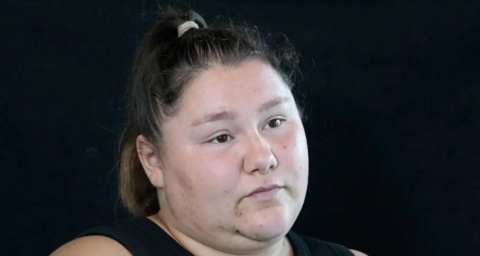 A woman with brown hair scraped back into a ponytail looks off camera in a head and shoulders image on a black background. She has brown eyes and is expressionless. She is wearing a black vest top.