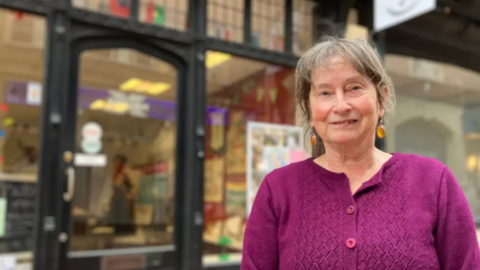 Sue Hagley with greying tied-back hair, amber dangly earrings and a purple cardigan outside the shop, which has black framed windows.