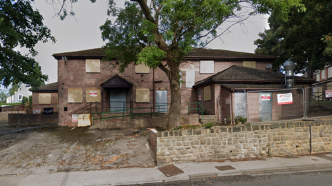 An abandoned building with some fencing around it, boarded up windows, and a few red and white safety precaution signs.