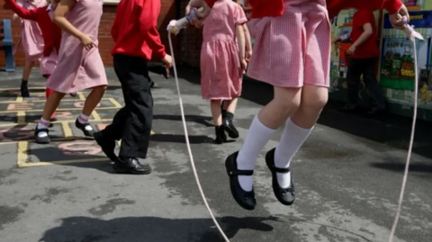 Girls and boys in playground jumping about and one person using a skipping rope
