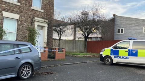 The house in Elswick where the attack took place. The property is an end terrace house made of brown brick. A climbing plant is growing on a trellis next to the white front door. Blue and white police tape has been strung from the property's wall and across a grey car. A police van is parked on the road nearby.