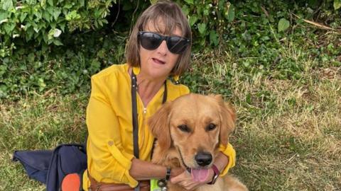 A woman in a yellow top cuddles a dog which has its tongue out
