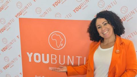 A woman with brown curly hair smiling at the camera. She is wearing an orange blazer over a white shirt. She is standing in front of an orange sign which she is gesturing towards.