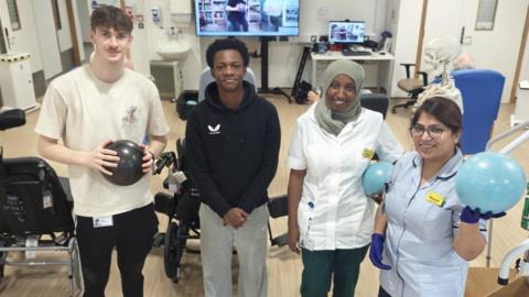 Two men and two women inside a hospital stroke recovery ward. There are wheelchairs behind them, along with physiotherapy exercise balls, computers and a large TV screen.