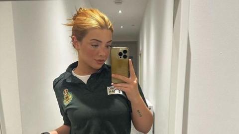 Lorna is using her mobile phone to take a selfie in a mirror here. She has blonde/ginger hair which is pinned up. She's wearing a green Welsh ambulance service polo shirt with a white shirt underneath.
