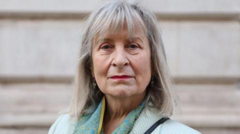A woman with shoulder length greying hair and a pale green jacket stares directly at the camera