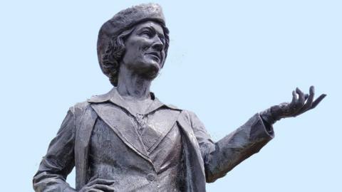 Close-up shot of a bronze statue of Nancy Astor, the first female MP to sit in the House of Commons. The statue shows Astor in a pose where her left arm is outstretched as she looks into the distance. It is a sunny day.