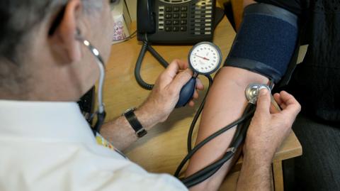 A doctor checking a patient's blood pressure with their arm leaning on a desk with a telephone on it.