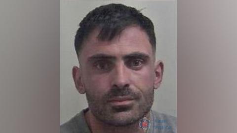 A custody image of Huseyin Kalyoncu. He has short brown hair and stubble. He is looking directly at the camera and is wearing a grey jumper.