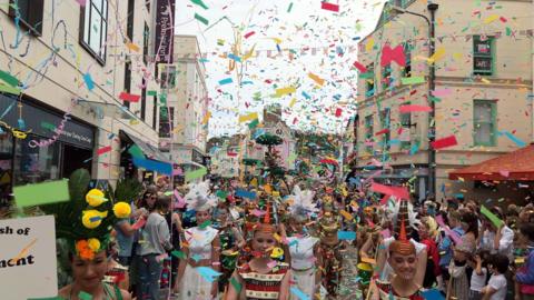 A street in St Helier engulfed by multicoloured confetti and people, some dressed in costume and others spectators of the Battle of Flowers.