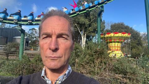 A man stands in front of a rollercoaster at a visitor attraction. You can see people on the amusement ride with their hands in the air. There is also a carousel in the background.