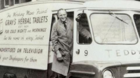 John Gray stands in the door of an old-fashioned van with The working man's friend Gray's Herbal Tablets for Cold Nights and Mornings written on the side. Gray is smiling and wearing a long apron, shirt and tie. 