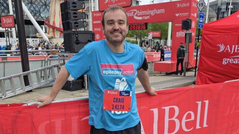 A man dressed in a blue sports t-shirt stands smiling at the side of the road with a medal around his neck