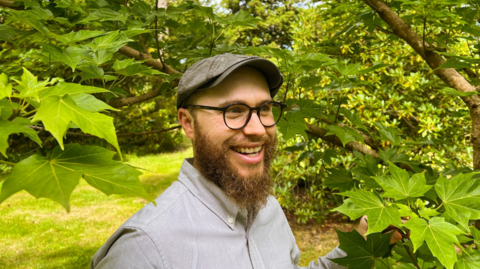 Dan Crowley standing among tree branches and leaves. He is wearing a flat cap, has dark-rimmed spectacles and a brown beard