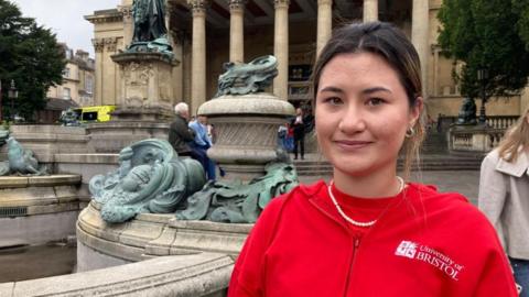 Yume Penwarden is pictured standing in front of the Victoria Rooms in Bristol. She is a young woman with dark hair that is tied back. She is wearing a bright red T-shirt underneath a red University of Bristol hoodie. She is smiling.
