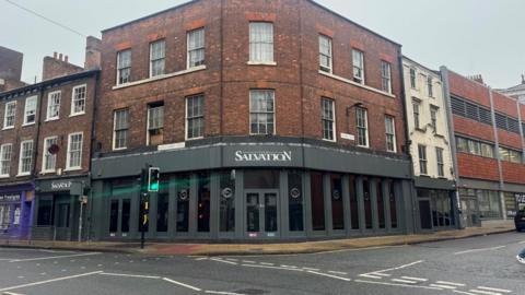 A three-story building on the corner of a asphalt road junction. The ground floor is grey painted window frames, the next two floors are red brickwork. White letters above a ground floor door entrance read Club Salvation.