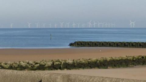 A view of a beach with sea defence and an offshore wind farm can be seen in the background