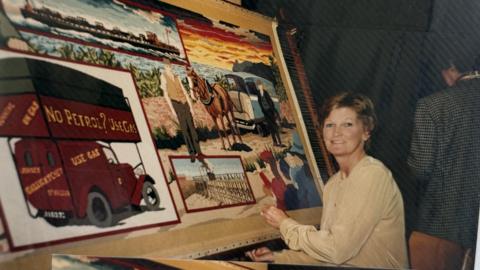 The picture shows a large, colourful textile artwork mounted on a wooden frame. The artwork appears to be a detailed tapestry panel depicting scenes from Jersey’s history, including a red vintage delivery van with the words “No Petrol? Use Gas” and “Jersey Gas” clearly visible, a horse pulling a cart in a rural setting, a background featuring fields, fences, and a vivid sunset sky with shades of orange, yellow, and purple.