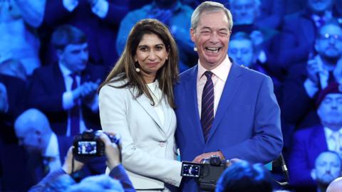 Suella Braverman and Nigel Farage with arms around each other smiling for cameras in front of a crowd. 