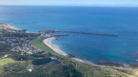 Alderney's Braye Harbour seen from the air with boats, the breakwater, beaches, houses and a lot of green space.