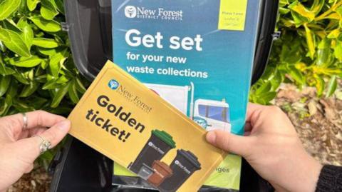 A piece of gold paper saying "golden ticket" on it, and an information leaflet about the new waste collections, being held up in front of a black bin.