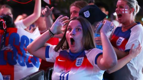 A woman in an England t-shirt looks shocked with her hands in the air while watching the Euro 2025 England v Italy semi-final at Wembley Boxpark, with people behind her shouting in celebration