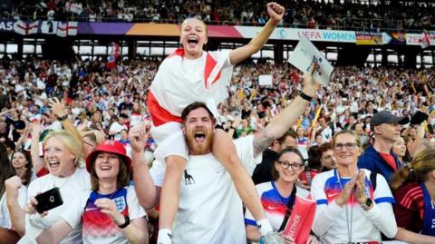 A young girl draped in an England flag sits on a man's shoulder surrounded by cheering England fans in a packed stadium