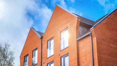 New apartments in a construction site. It's a stock image and the building is red brick with grey window fixings. There are also clear half barriers over the windows. 