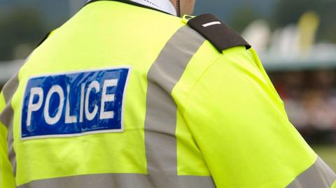 The back of a police officer who is wearing a high-viz yellow jacket with grey lining. The word POLICE is spelled out in white letters on a blue background on a panel towards the top of his back
