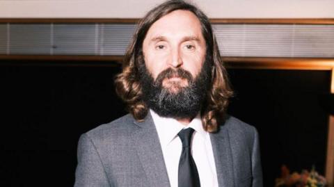 A bearded man, with shoulder-length brown hair looks at the camera. He is wearing a grey suit, white shirt and a black tie