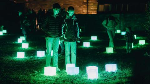 A series of light boxes in a field. Surrounding them are different people looking at them.