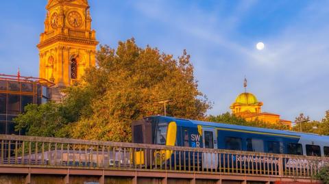 The sun rises over Portsmouth with a blue and yellow train passing over a bridge in the foreground. Behind there is a stone clock tower glowing yellow in the sun and a green tree. The sky is blue with wisps of white cloud. 