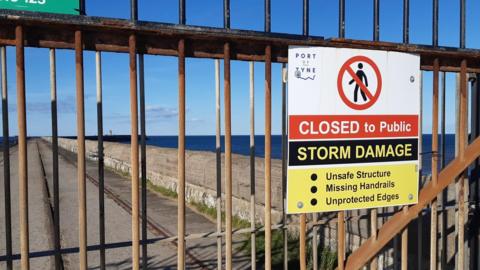A rusted metal gate at the entrance of the South Pier at South Shields. A sign attached has the outline of a person crossed out. It reads: "Closed to public. Storm damage".