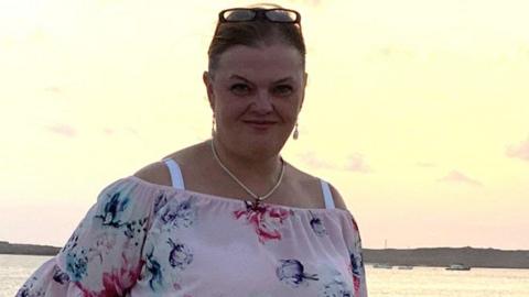 A woman, with sunglasses on her head, with a necklace round her neck, and earrings in her ears, with dark hair put up, by water, and wearing a white floral top. She is looking at the camera.