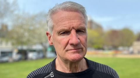 Deputy Tom Binet wearing a black top and looking at the camera. He is stood in a green park which is blurred by the camera.