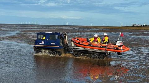 A blue vehicle tows an orange lifeboat with a small crew on board across the wet sands of the Dee Estuary. Wind turbines can be seen in the far background.