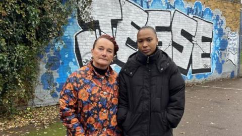 Sam Wellsted, Mr Lloyd-Smith's godmother (left), standing next to his sister Chaise Lloyd-Smith. Ms Wellsted is  a woman with red hair wearing a floral coat, and Ms Lloyd-Smith is a woman with dark hair wearing a black padded coat. They stand in front of a blue and white spray-painted mural saying the name Jesse