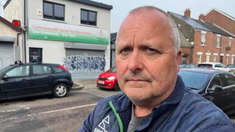 Conal Heatley is staring into the camera, wearing a blue jacket and grey T-Shirt. He is standing in front of the Commonewealth Games Northern Ireland offices, which are shut