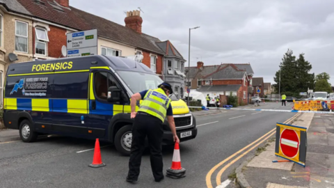 A police van with the word "forensics" on it. A police officer is laying out cones nearby.
