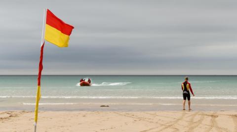 A beach showing a flag marking the safe swimming area. A lifeguard is stood on the beach while looking at a rescue boat heading towards shore. 