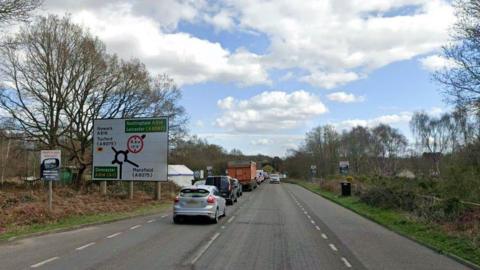 A street view of the A614 in Nottinghamshire showing a road sign for a roundabout on the left of the road. A number of cars are queuing and there are trees lining the road on both sides. 