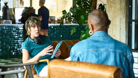 Young man in green T -shirt with headphones around hs neck having a serious discussion with a woman in denim seen from behind. They are in a modern contemporary cafe setting
