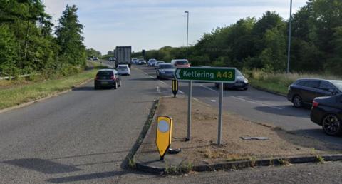 Road with single carriageway on either side with a lot of traffic and a sign pointing to Kettering