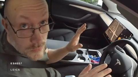 A bald man wearing glasses and with a grey and brown beard. He is sat in the driver's seat of a Tesla car. It is captured on bodyworn camera as he is being arrested